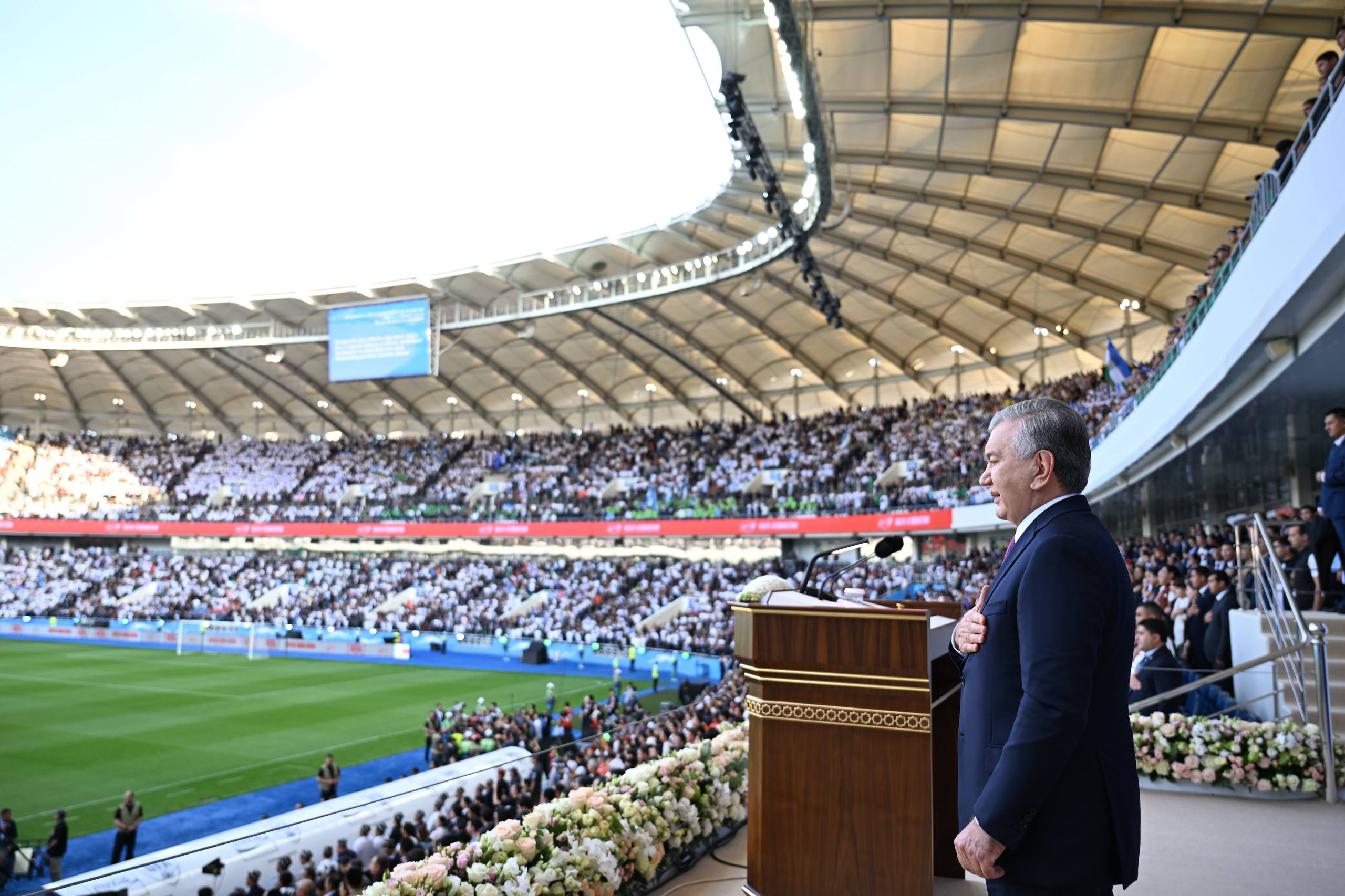 Shavkat Mirziyoyev gavjum stadionda qo‘lini ko‘ksiga qo‘yib turibdi.