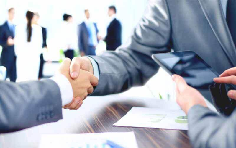 Two people in suits shaking hands over a table.