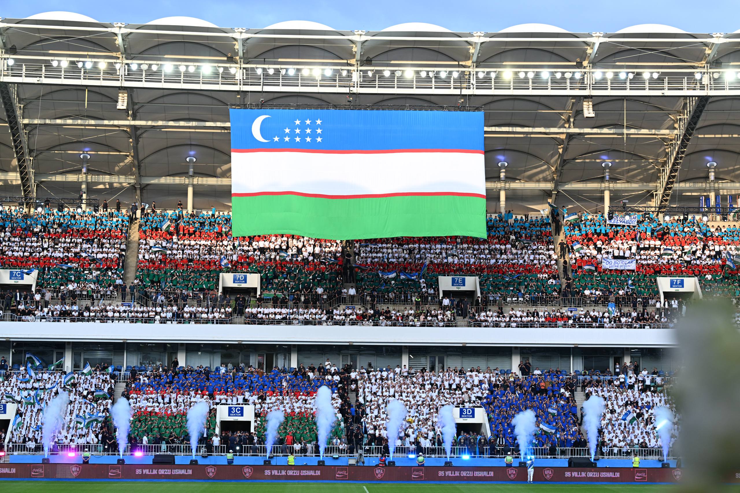 Stadionda O‘zbekiston bayrog‘i ostida yig‘ilgan ko‘p sonli muxlislar va tutun effektlari.