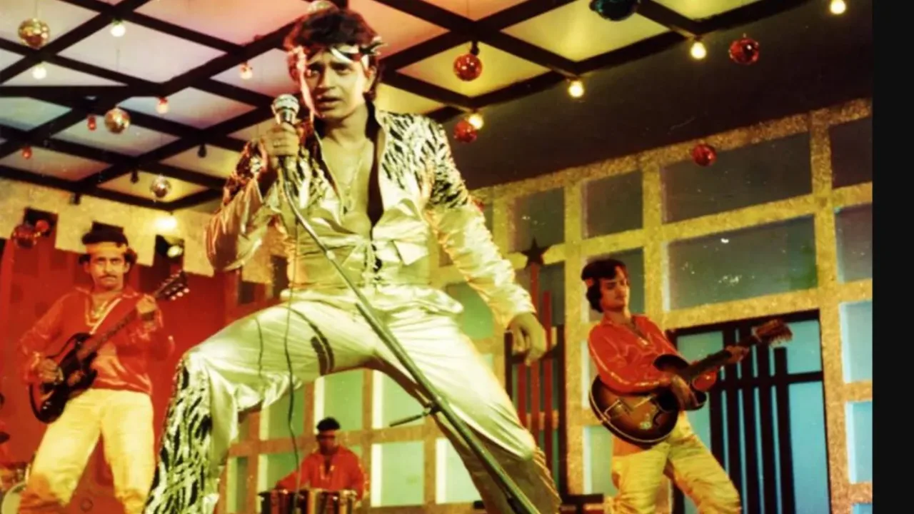 Mithun Chakraborti yaltiroq kiyimda sahnada qo‘shiq kuylamoqda.