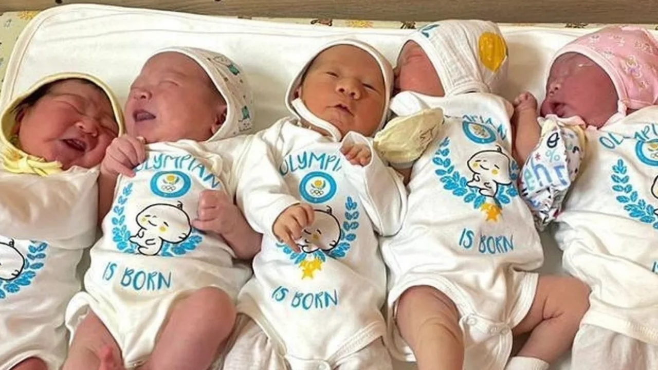 Olimpiya ramzi tushirilgan oq kiyimlardagi besh nafar yangi tug‘ilgan chaqaloq yonma-yon yotibdi.