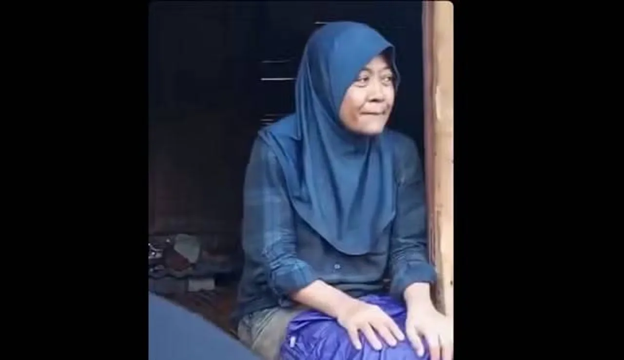 Ko'k rangli hijob va katakli ko'ylak kiygan ayol eshik oldida o'tiribdi.