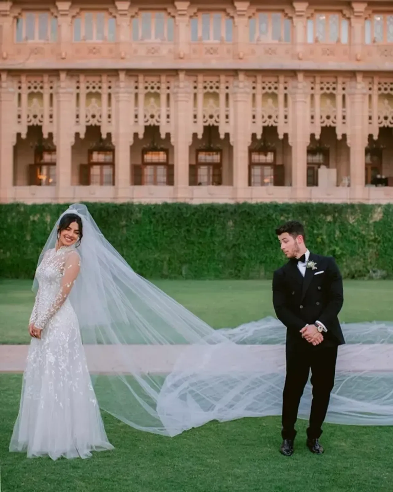 Priyanka Chopra and Nick Jonas standing in wedding attire in front of a luxurious building.