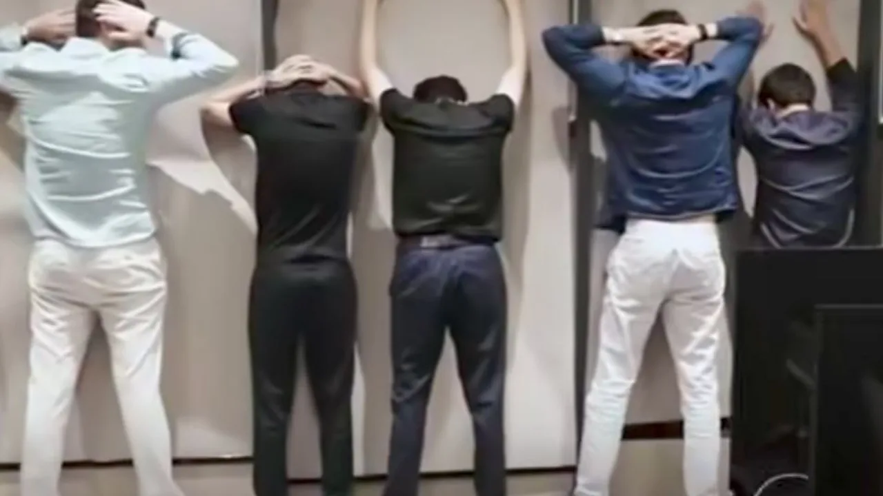 Five men are standing with their hands on their heads or leaning against a wall, viewed from behind.