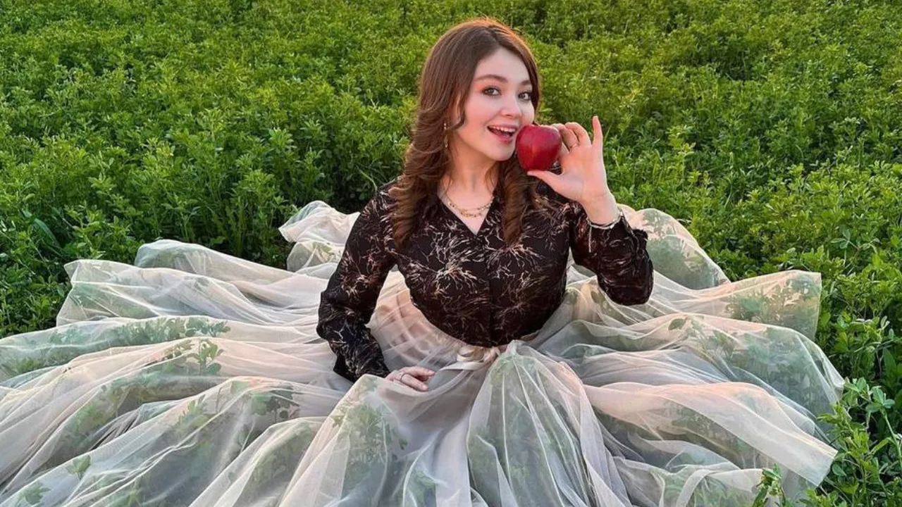 Keng ko‘ylak kiygan ayol yashil maydonda qizil olma ushlab o‘tiribdi.