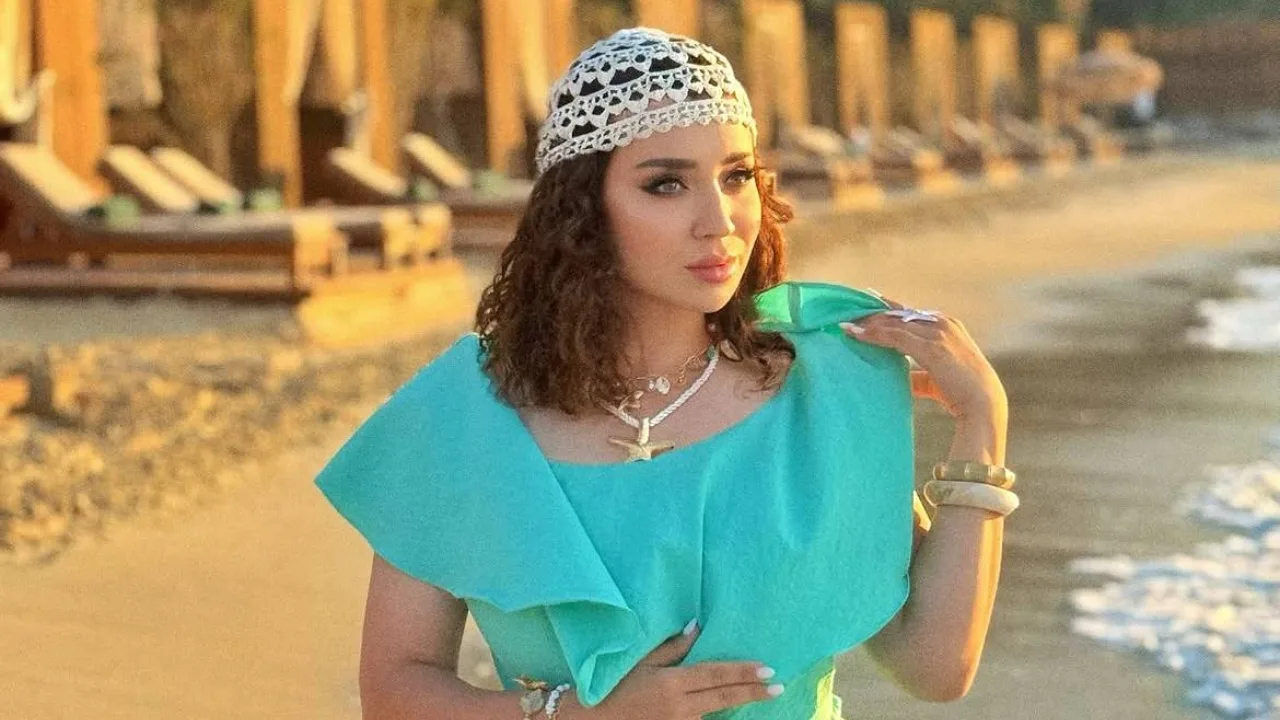 Firuza rang ko'ylak va oq to'rli telpakdagi ayol qumli sohilda turibdi.