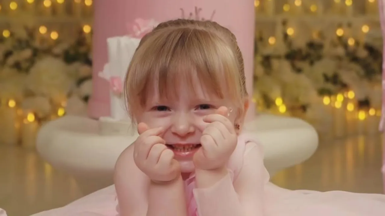 A little girl in a pink dress smiling and making a heart shape with her fingers.