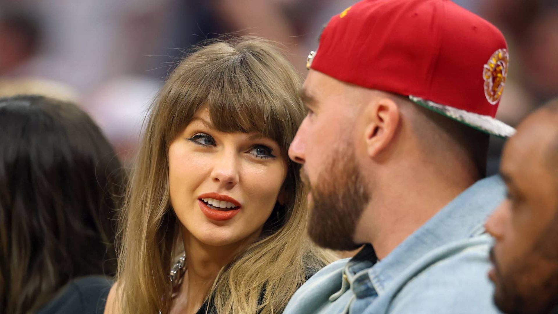 Taylor Swift smiling at Travis Kelce, who is wearing a red cap.