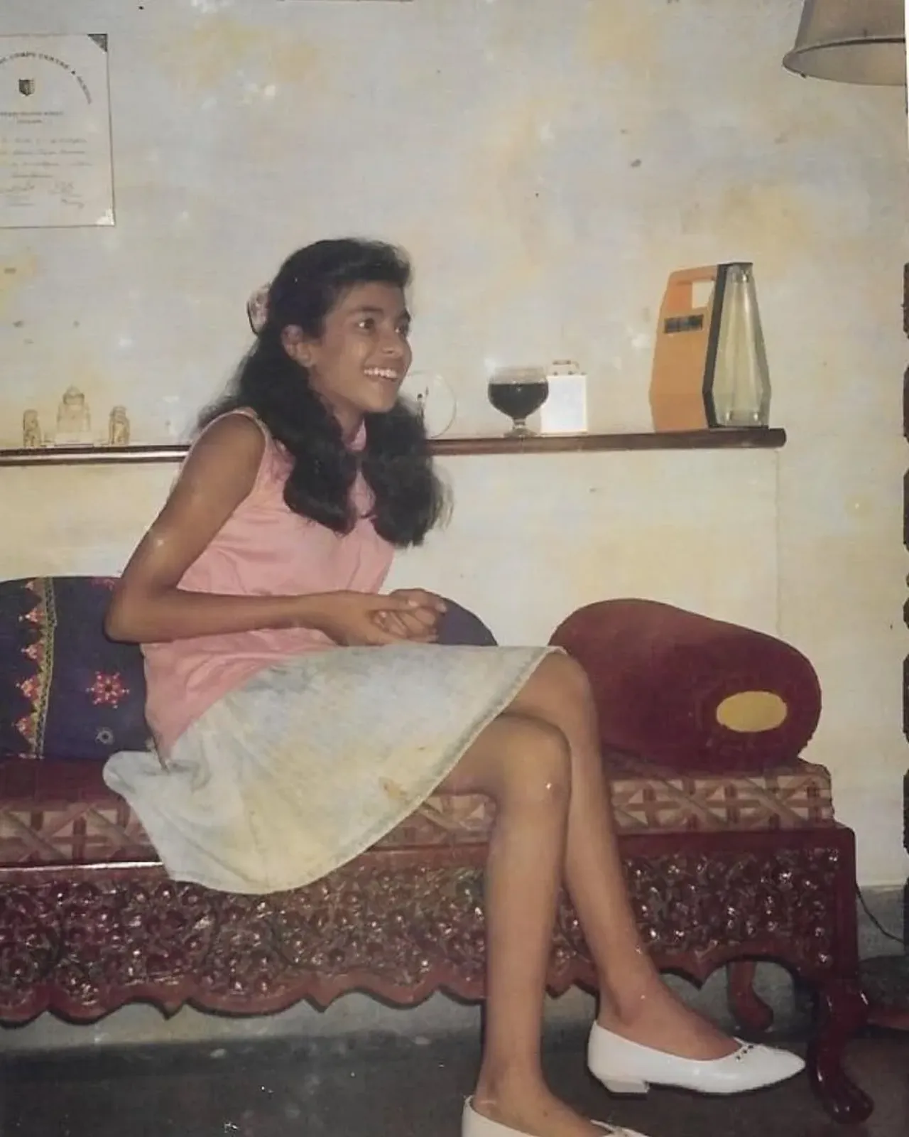 A young girl in a pink top and white skirt sitting smiling on a carved sofa.