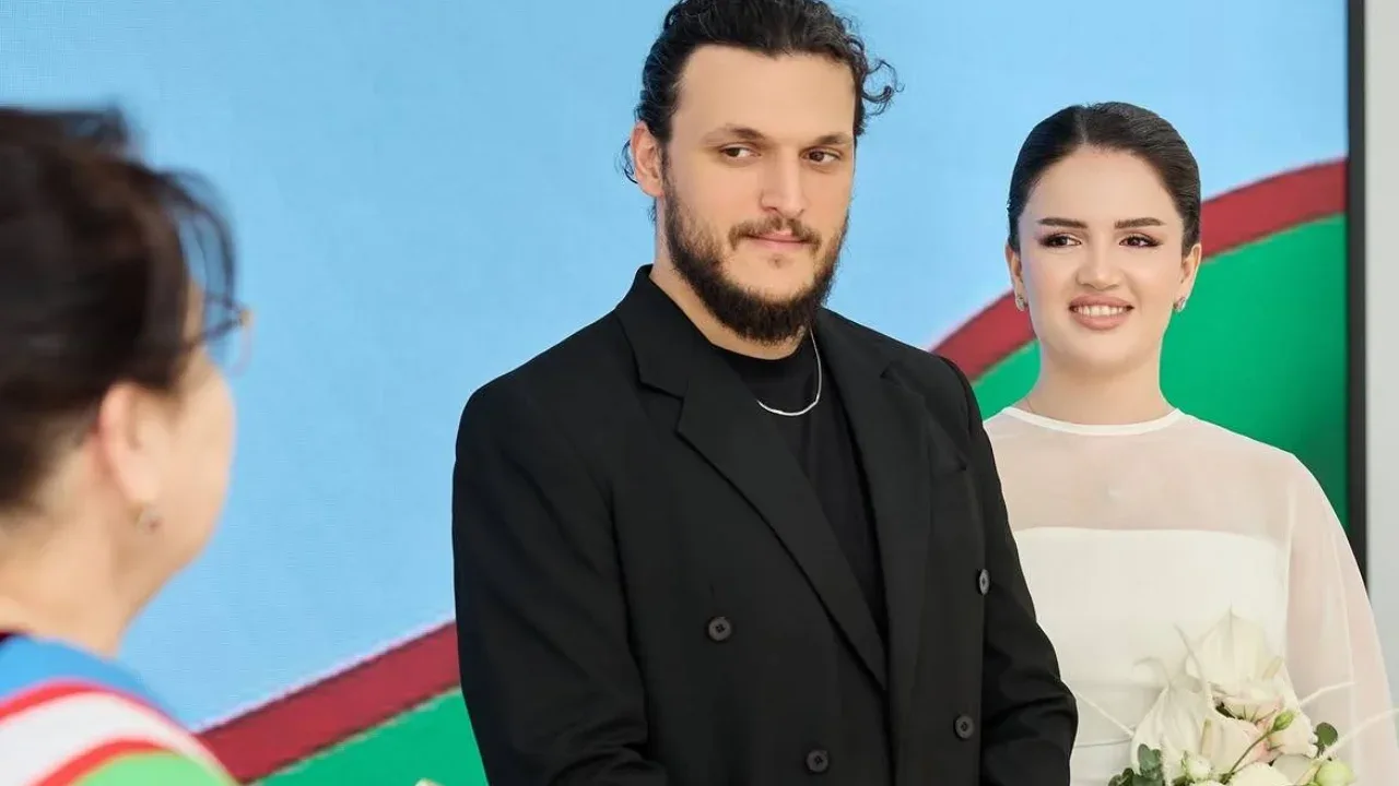 A bearded man in a black suit and a woman in a white dress are standing together.
