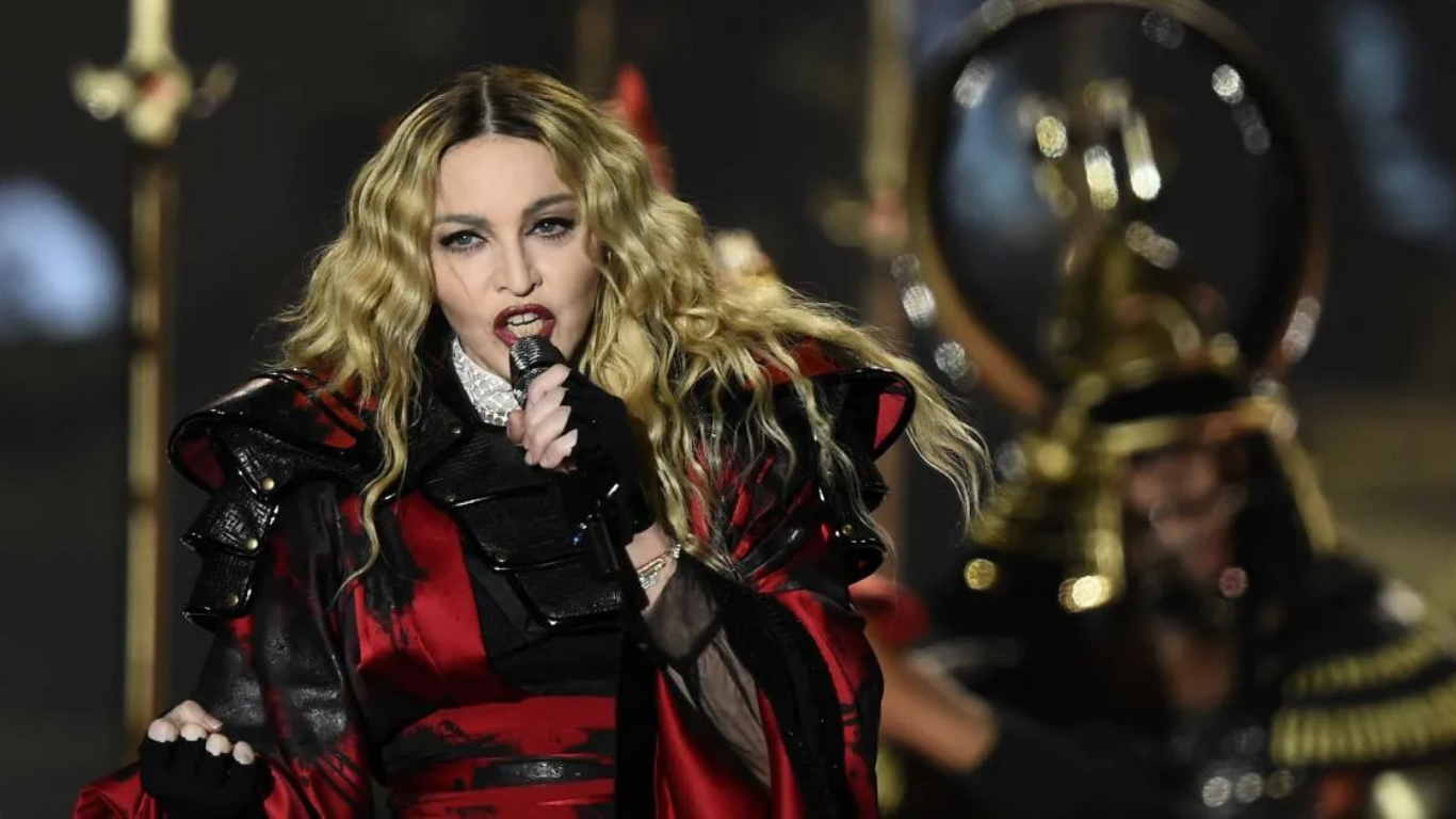 Madonna qizil va qora libosda sahnada mikrofon bilan qo‘shiq kuylamoqda.