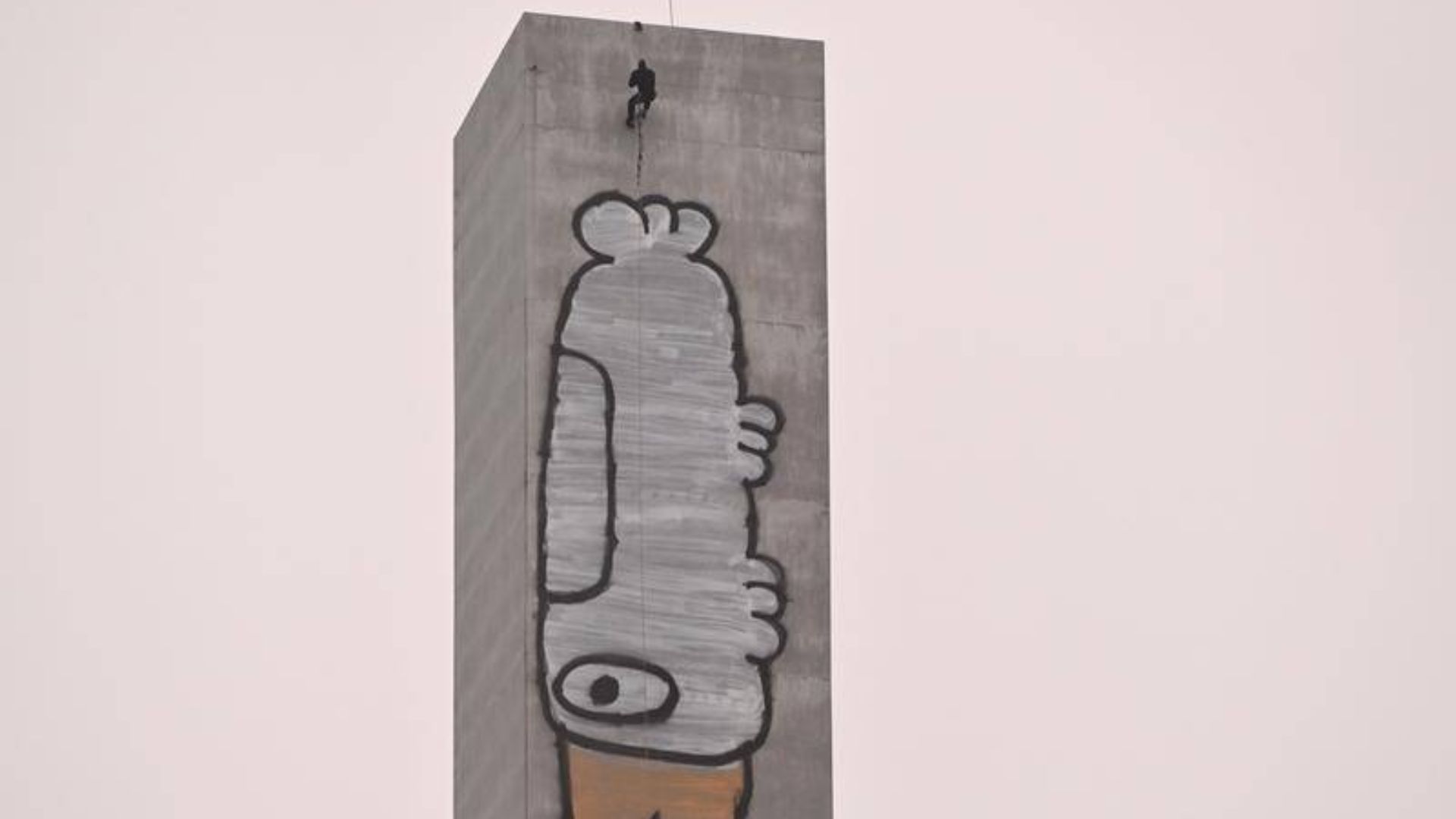 Baland beton binoning tepasida odam arqon bilan osilib turibdi va devorga graffiti chizmoqda.