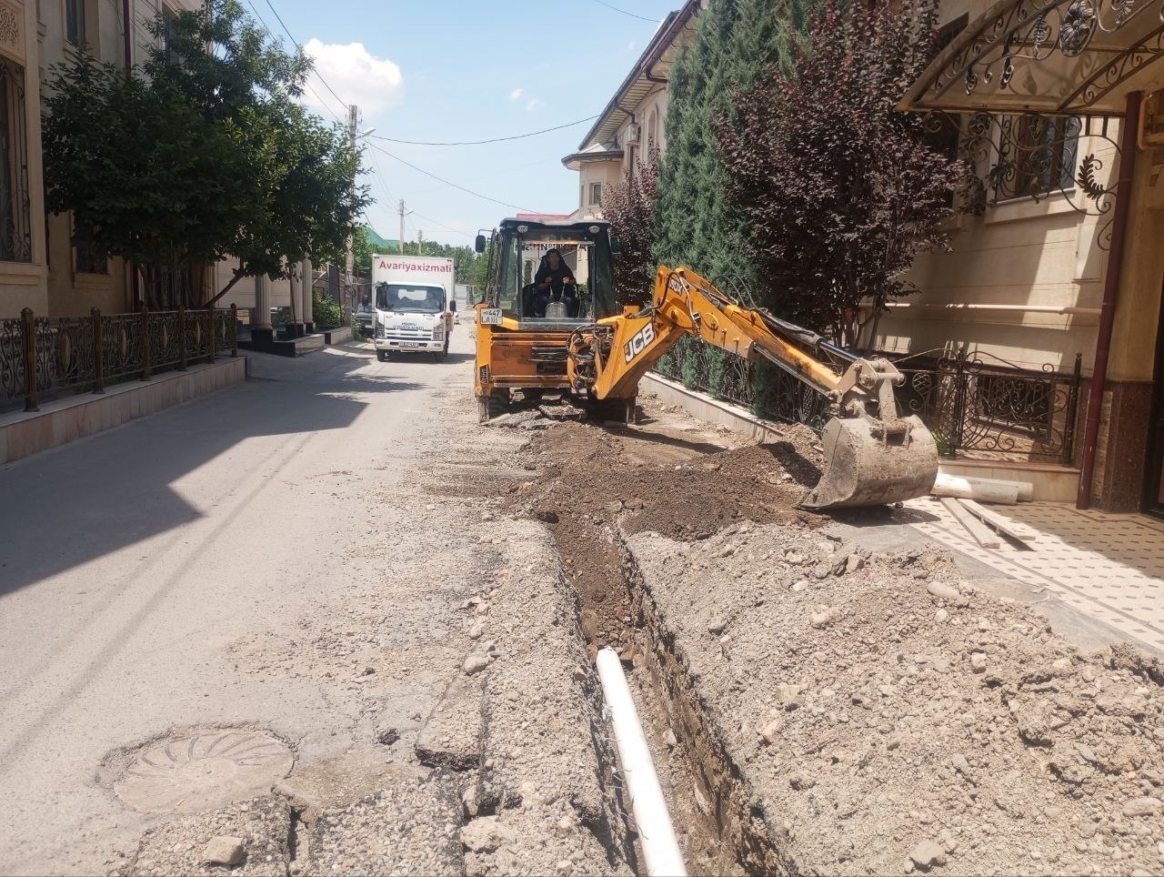 Sariq ekskavator tor ko‘chada quvur yotqizish uchun yer qazimoqda.