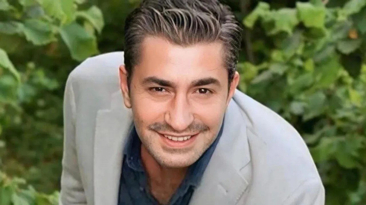 Yashil barglar fonida kulrang kostyumdagi jilmayib turgan erkakning portreti.