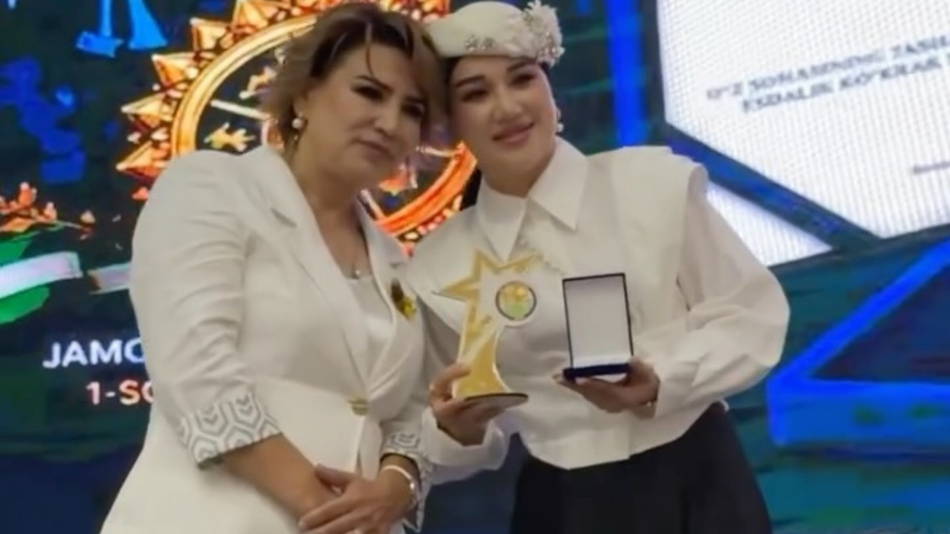 Two women in white clothes standing on stage with a star-shaped award and a medal.