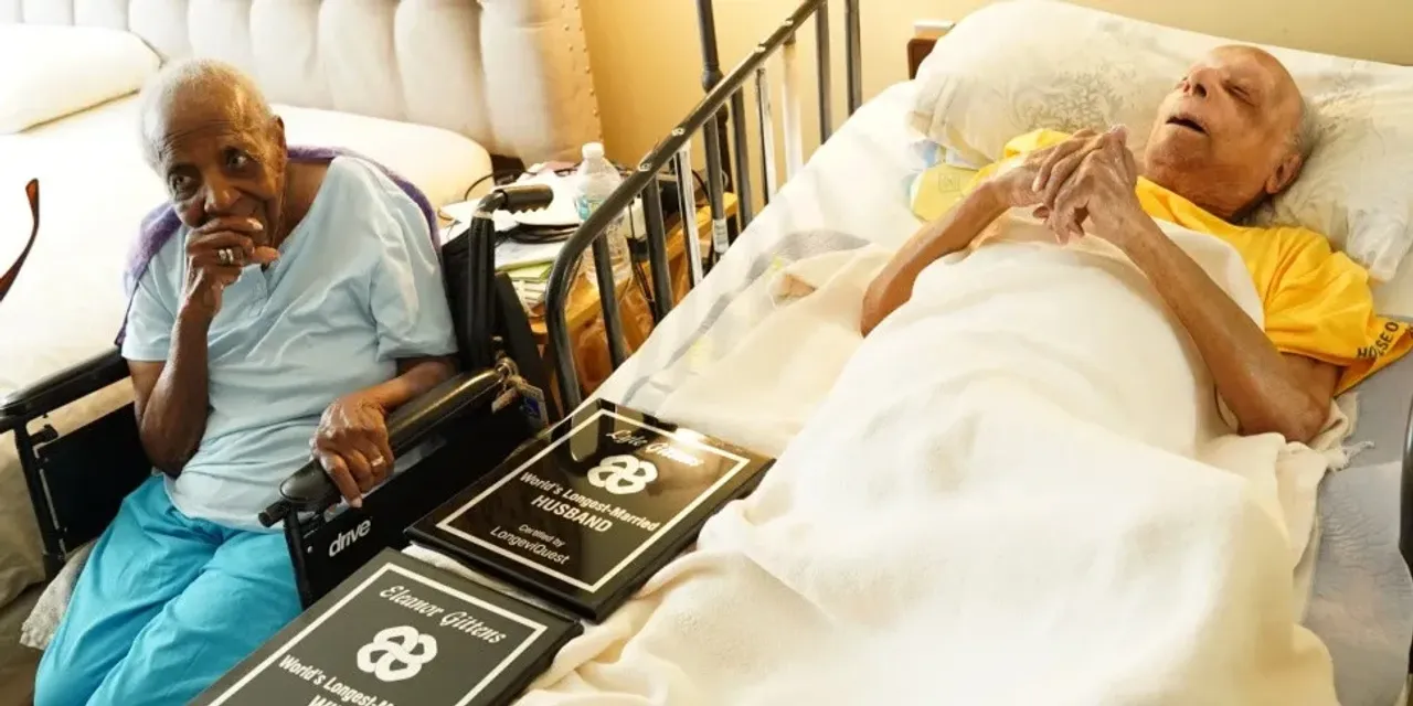 An elderly woman in a wheelchair, while an elderly man lies in bed.