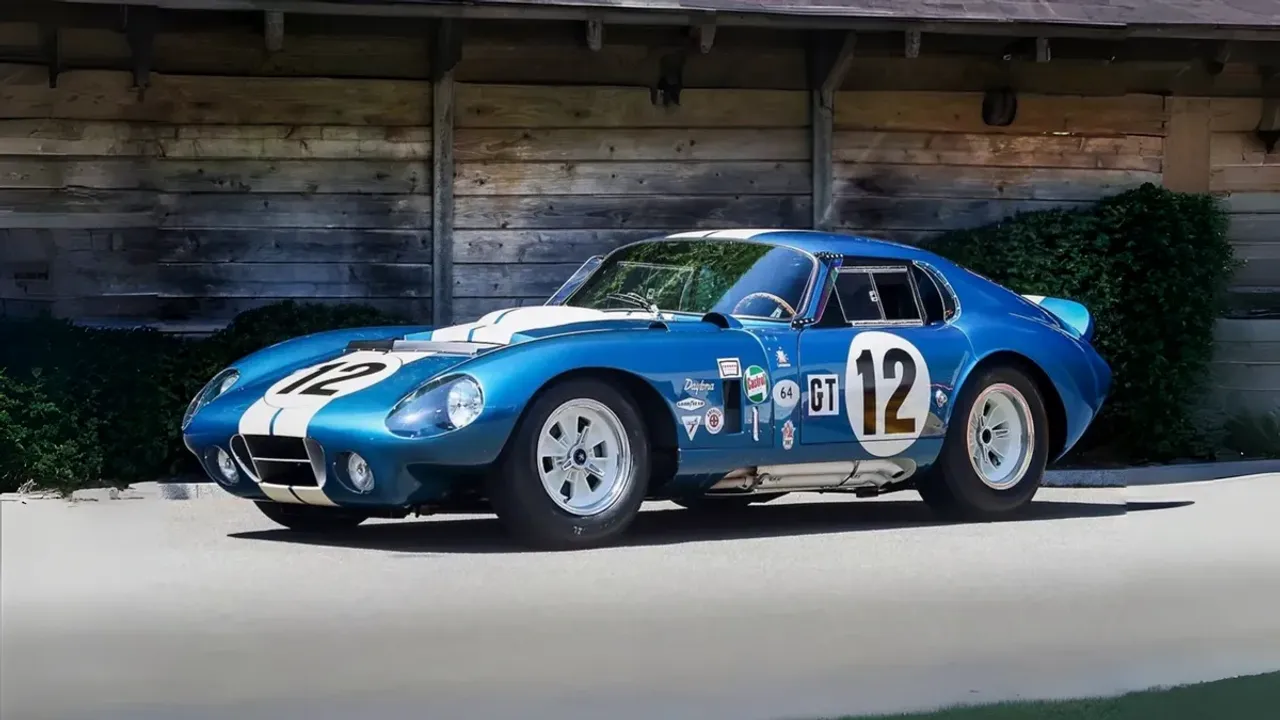 Yog‘och bino oldida 12 raqamli ko‘k Shelby Daytona Coupe poyga avtomobili turibdi.