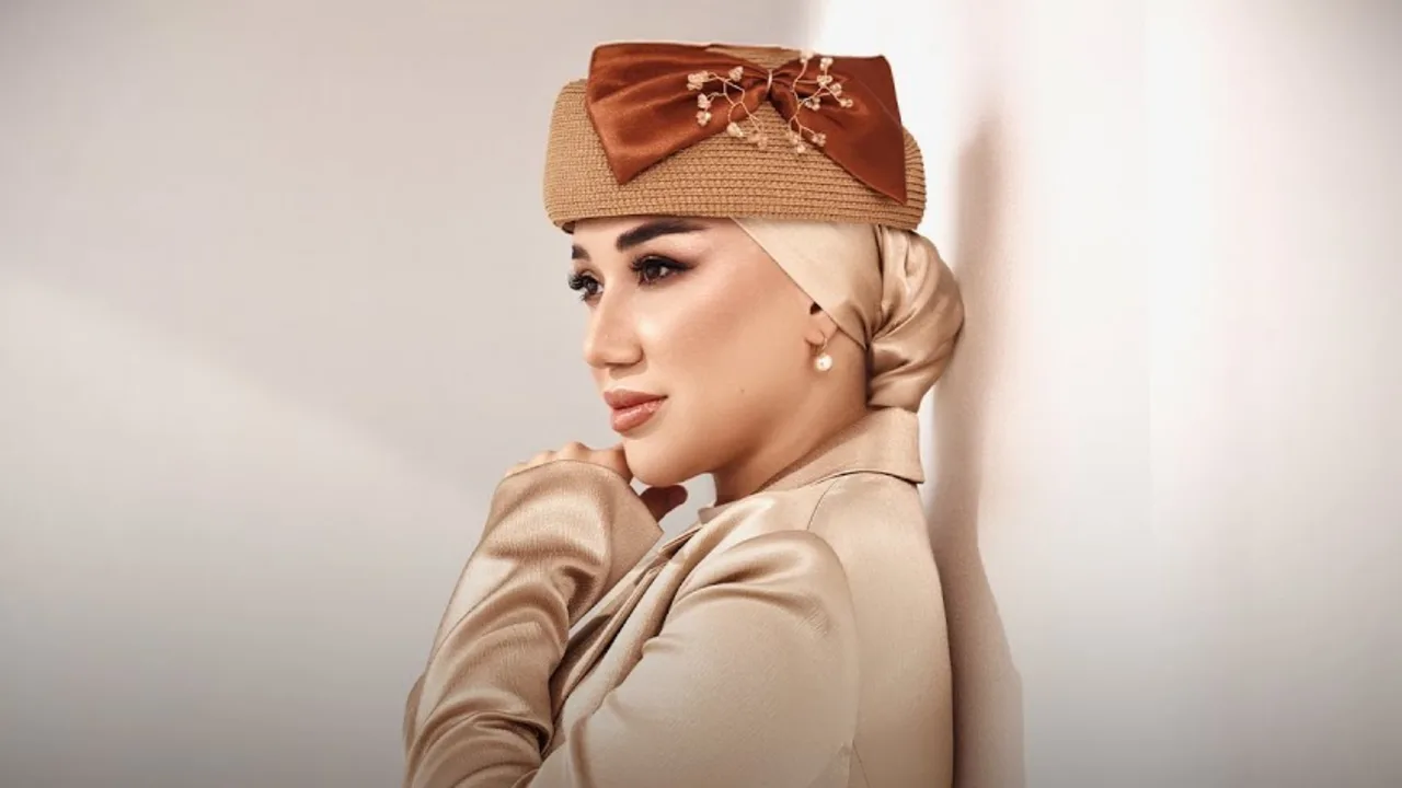 Jigarrang bantli shlyapa va ipak ro‘mol kiygan ayol yon tomonga qarab turibdi.