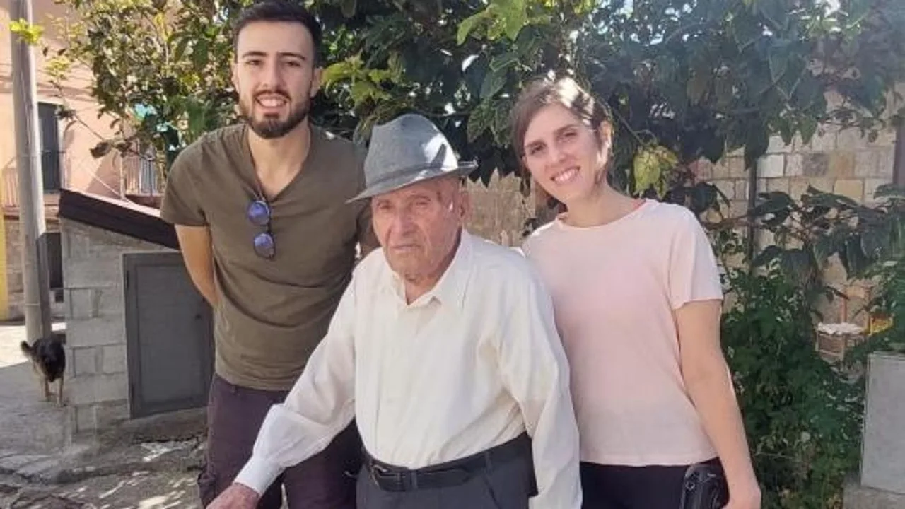 Ikki yosh kishi shlyapa kiygan keksaning yonida tabassum bilan turibdi.