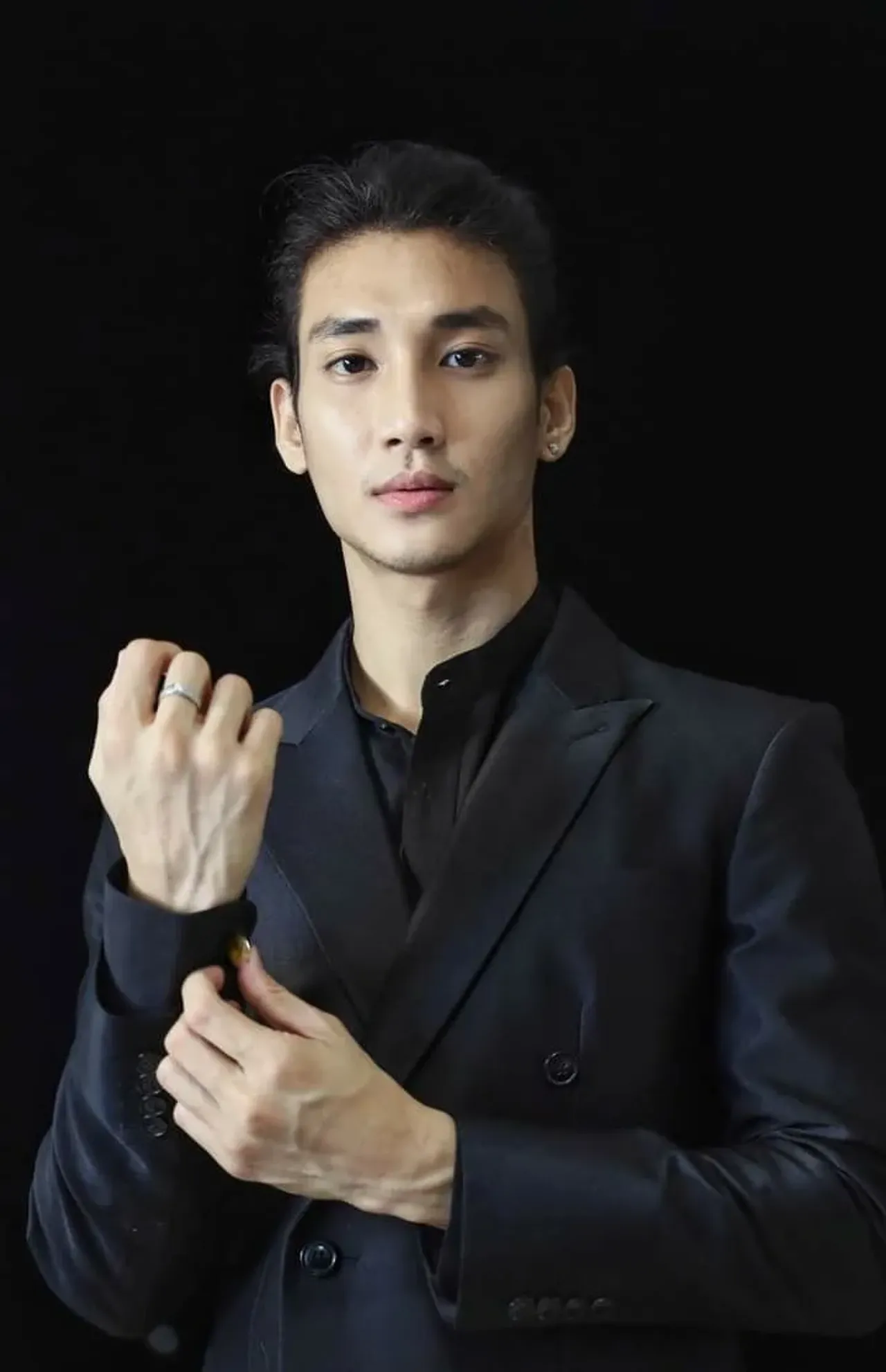 A guy in a black suit is fixing his buttons, standing against a black background.