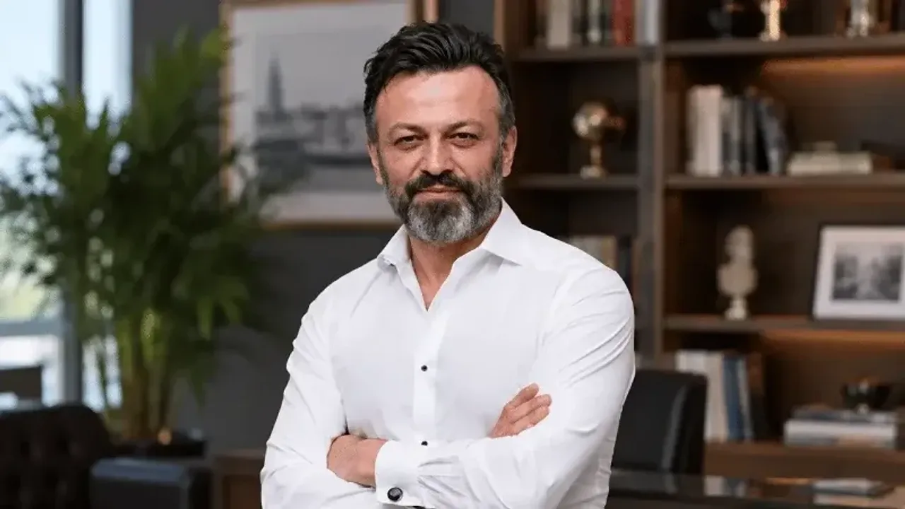 Oq ko'ylakli, soqolli kishi ofisda qo'llarini chatishtirib turibdi.