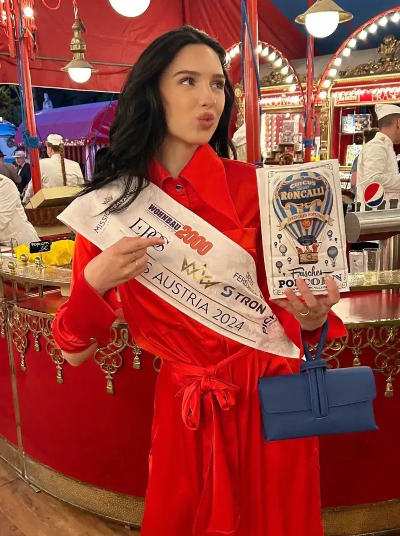 Qizil ko'ylakdagi Miss Avstriya 2024 g'olibi sirkda popkorn paketi bilan turibdi.