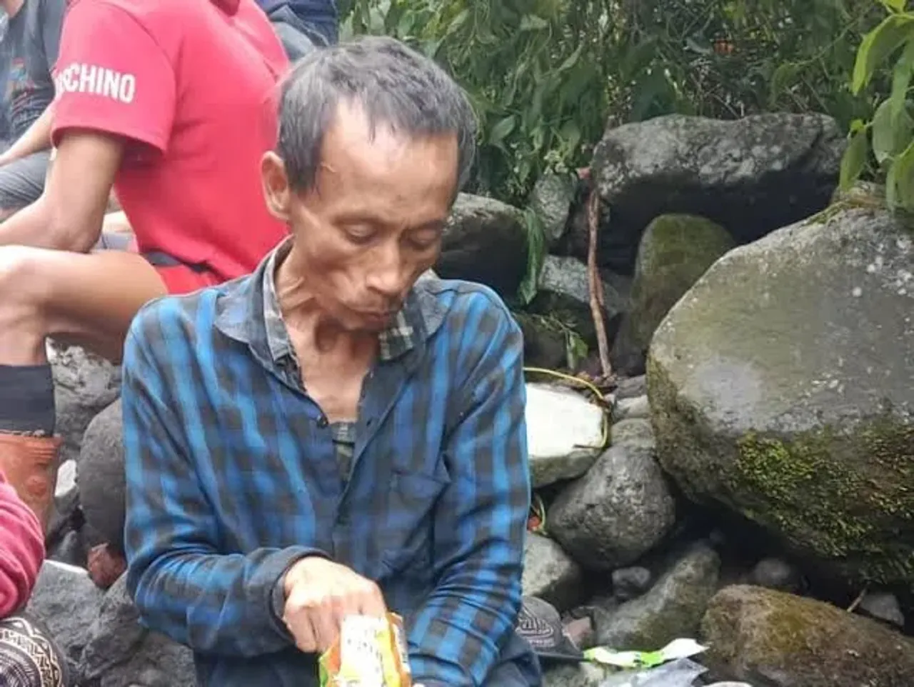 Katak-katak ko'ylak kiygan yoshi katta kishi toshlar oldida ovqat yemoqda.