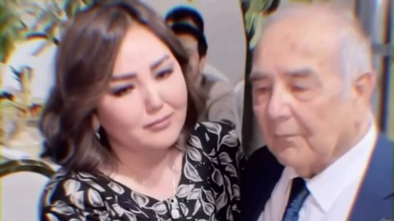 Gul kiyimdagi ayol va kostyum-shim kiygan keksa erkak yonma-yon turibdi.