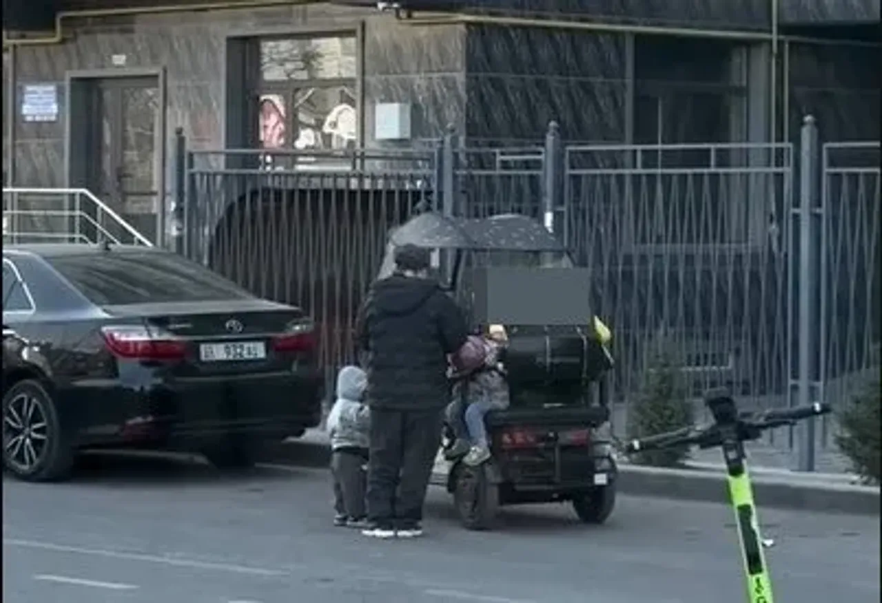Человек в черной одежде стоит на улице рядом с детьми и мопедом.