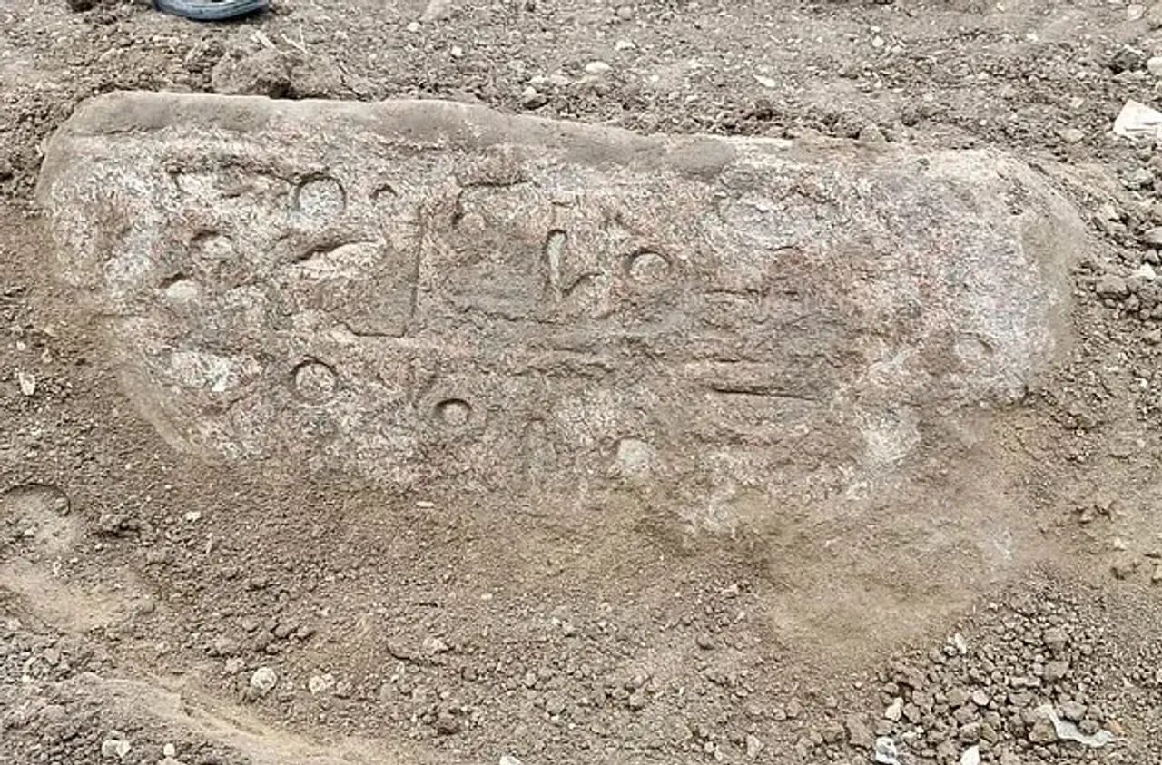 Yuzasida qadimiy yozuvlar va belgilar o‘yilgan yirik tosh tuproq ustida yotibdi.