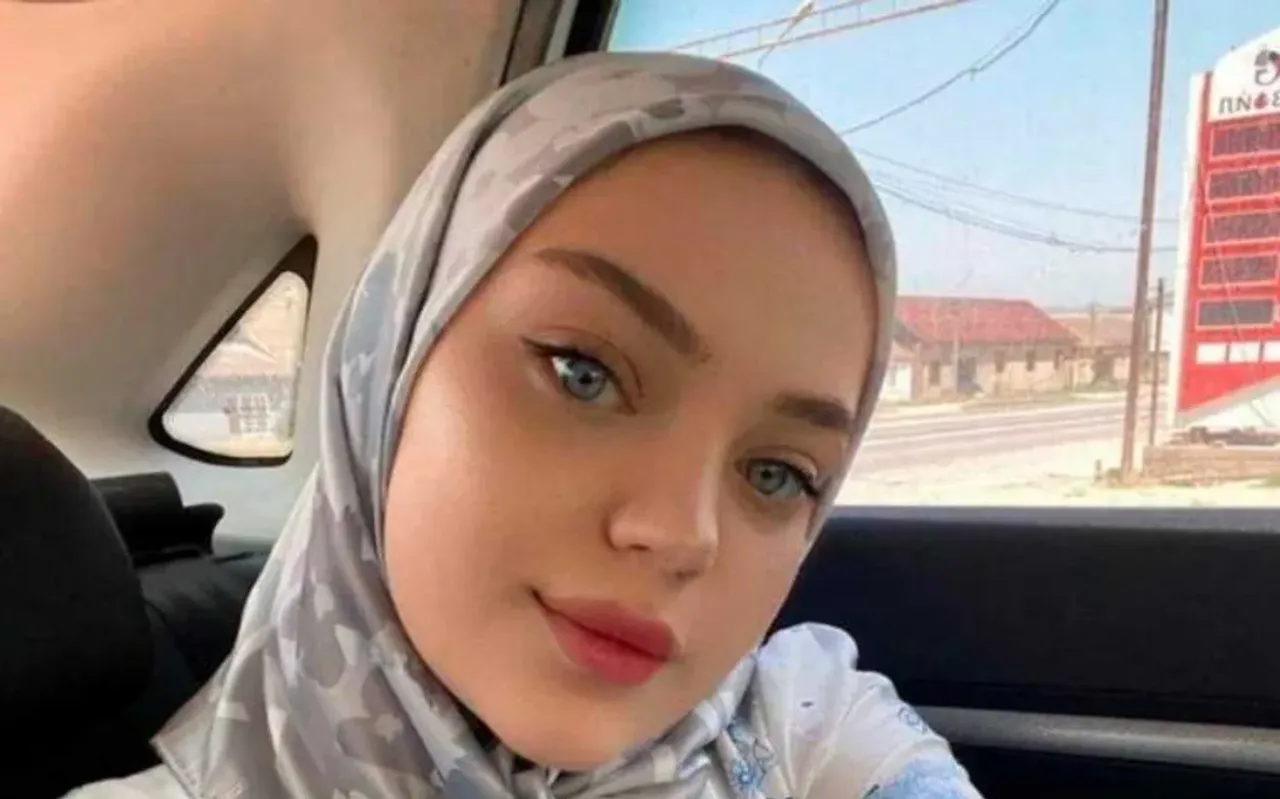 Avtomobil ichida kulrang ro'mol o'ragan moviy ko'zli yosh ayol selfi tushmoqda.