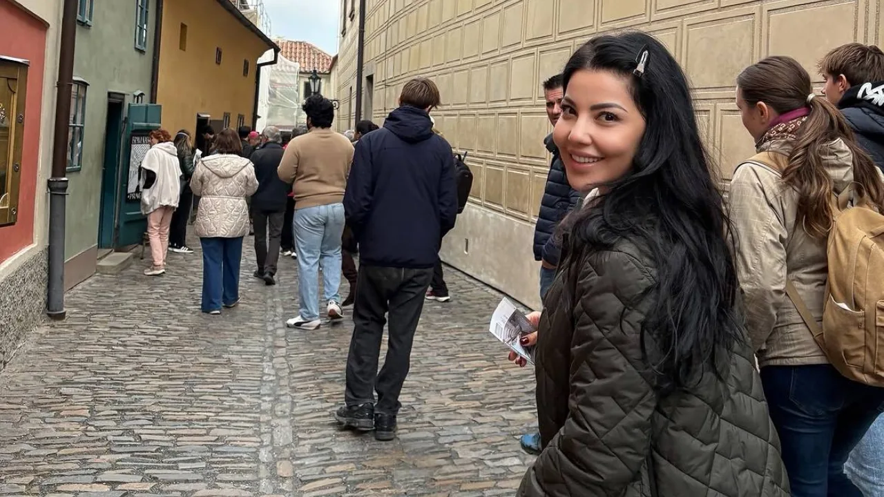 Qora sochli ayol toshli tor ko'chada odamlar orasida jilmayib turibdi.